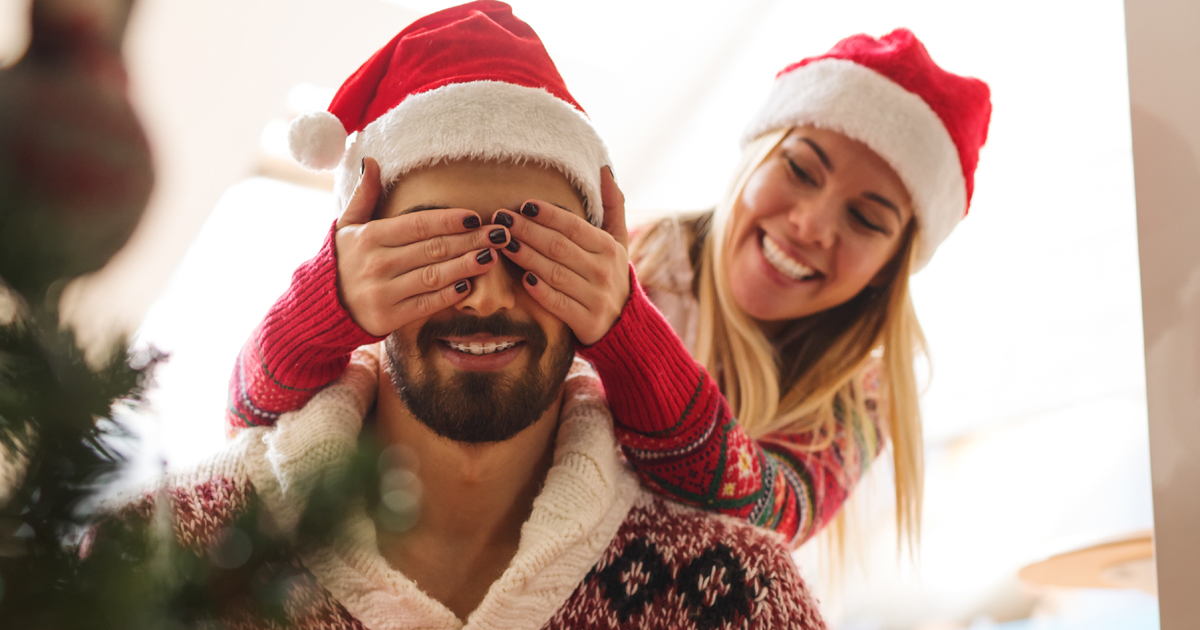 A woman covering her partner's eyes by a Christmas tree